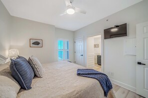 Een slaapkamer met een bed, kussens, een plafondventilator, een televisie en een deur naar een badkamer.