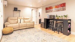 Una sala de estar con un sofá color beige, una mesa de centro y un tapete estampado. Se observa una cocina con un taburete de bar y un refrigerador.