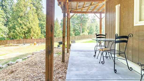Un porche cubierto con piso de concreto, sillas metálicas y una pérgola de madera.