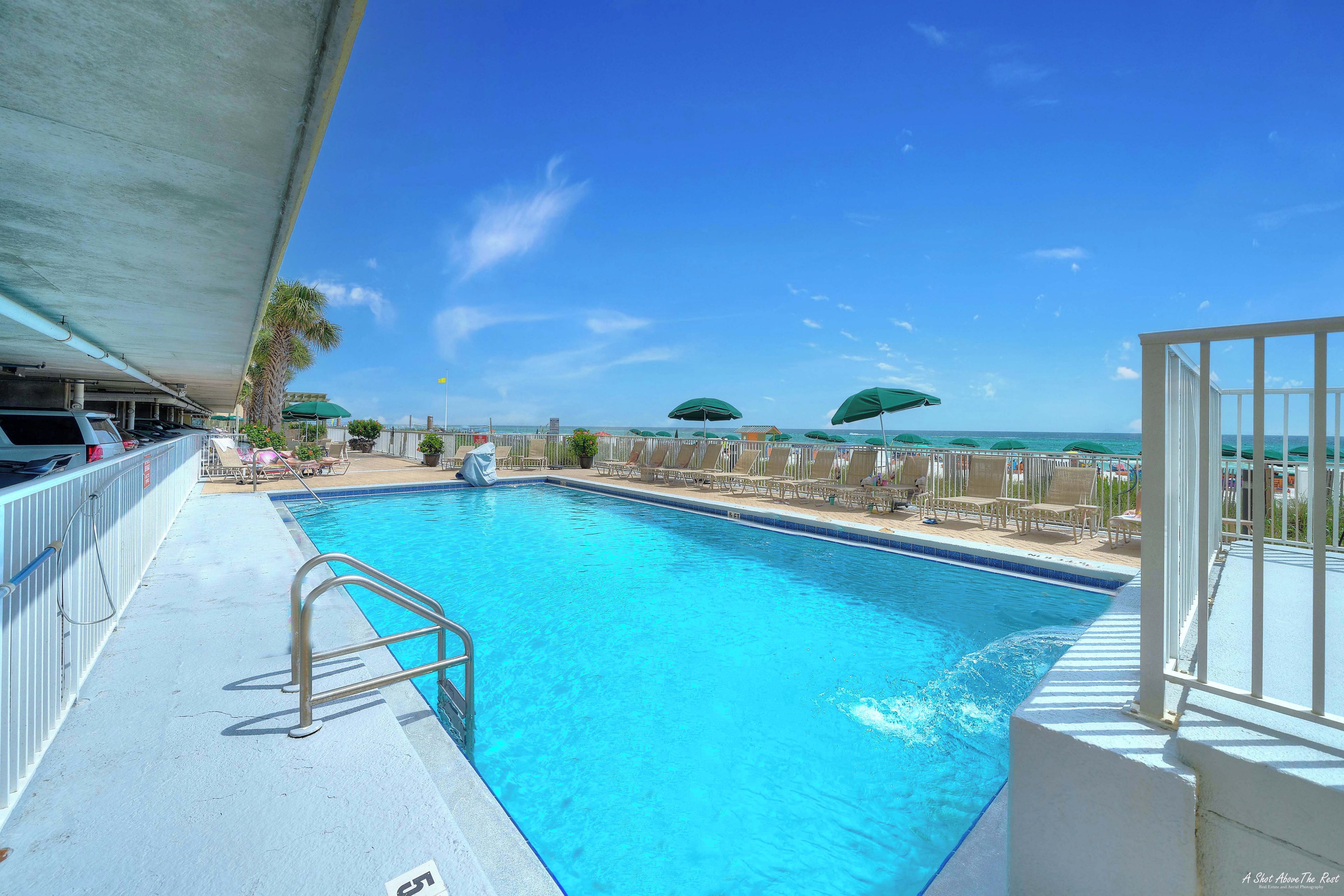 A swimming pool with a metal ladder, surrounded by lounge chairs and umbrellas, with a view of the ocean.