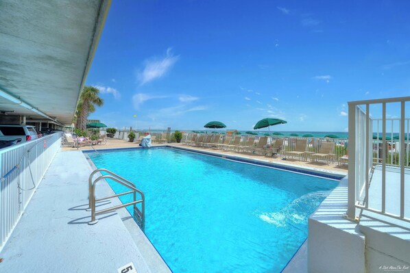 A swimming pool with a metal ladder, surrounded by lounge chairs and umbrellas, with a view of the ocean.