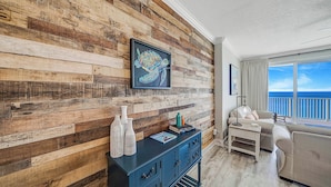A room with a wooden wall, a framed artwork, a blue cabinet, a white sofa, and a view of the ocean. - NEW Emerald Isle 2107 "Manta Ray Villa" Pool, FREE Perks&Beach Service for 4! (Panama City Beach)