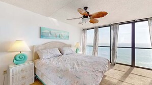 Een slaapkamer met een groot bed, een plafondventilator, een nachtkastje met lamp en uitzicht op de oceaan door grote ramen.