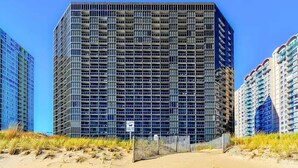 Een hoog gebouw met een geavanceerde gevel, omringd door duinen en andere hoge gebouwen.