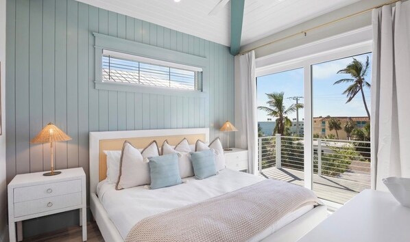 A bedroom with a bed, nightstands, a window with blinds, and a balcony with a view of palm trees and buildings.