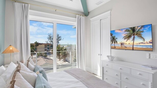A bedroom with a bed, a nightstand, a TV, and a view of the beach through the window.