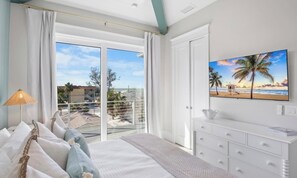 Ein Schlafzimmer mit einem Bett, einem Nachttisch, einem Fernseher und Blick auf den Strand durch das Fenster.