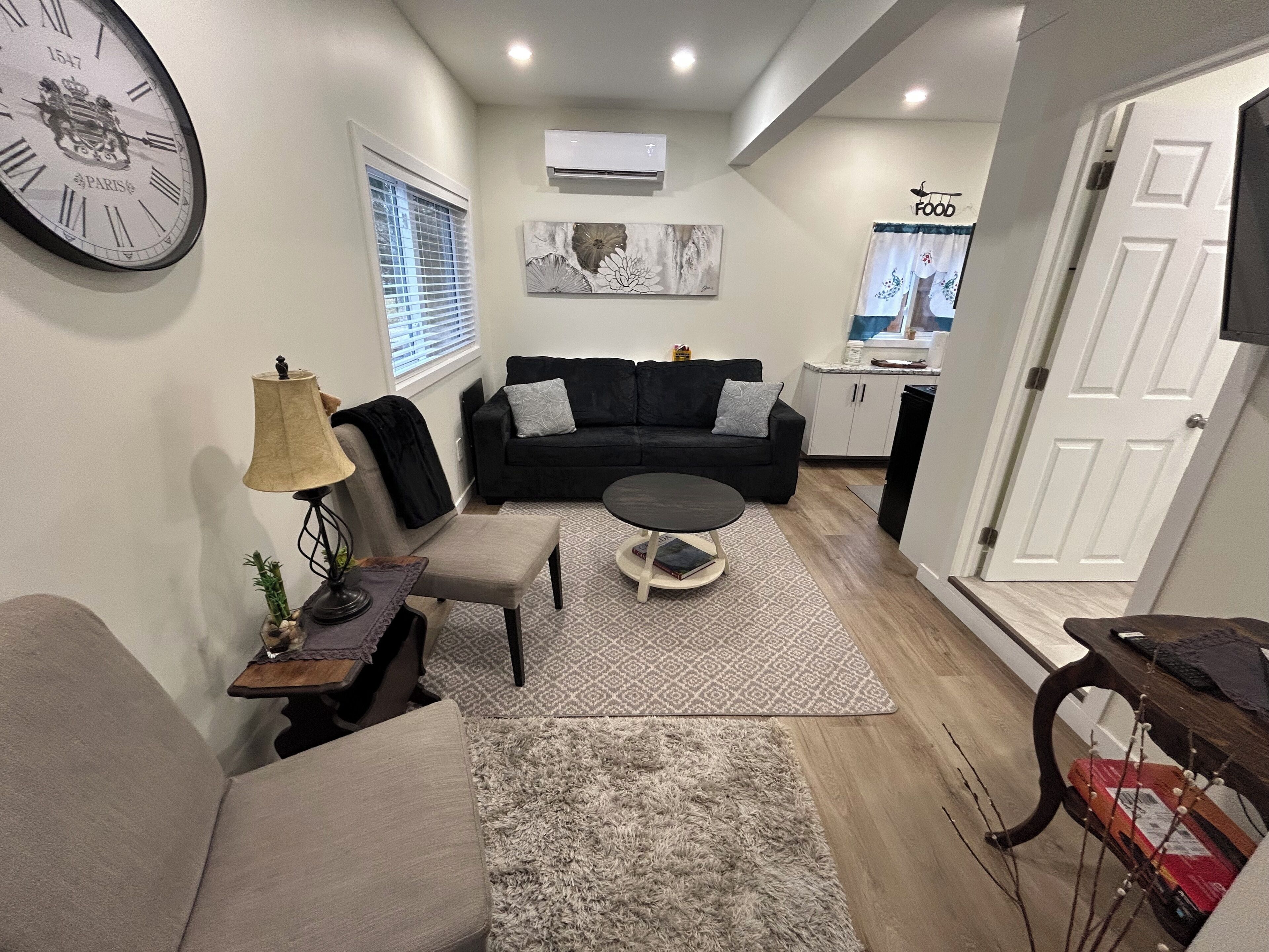 A living room with a black sofa, a round coffee table, a grey armchair, a lamp, and a clock on the wall.
