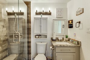 A bathroom with a glass-enclosed shower, a toilet, a sink with a marble countertop, and a vanity with drawers.