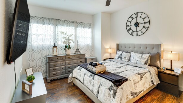 A bedroom with a bed, a ceiling fan, a clock, a dresser, and a television.