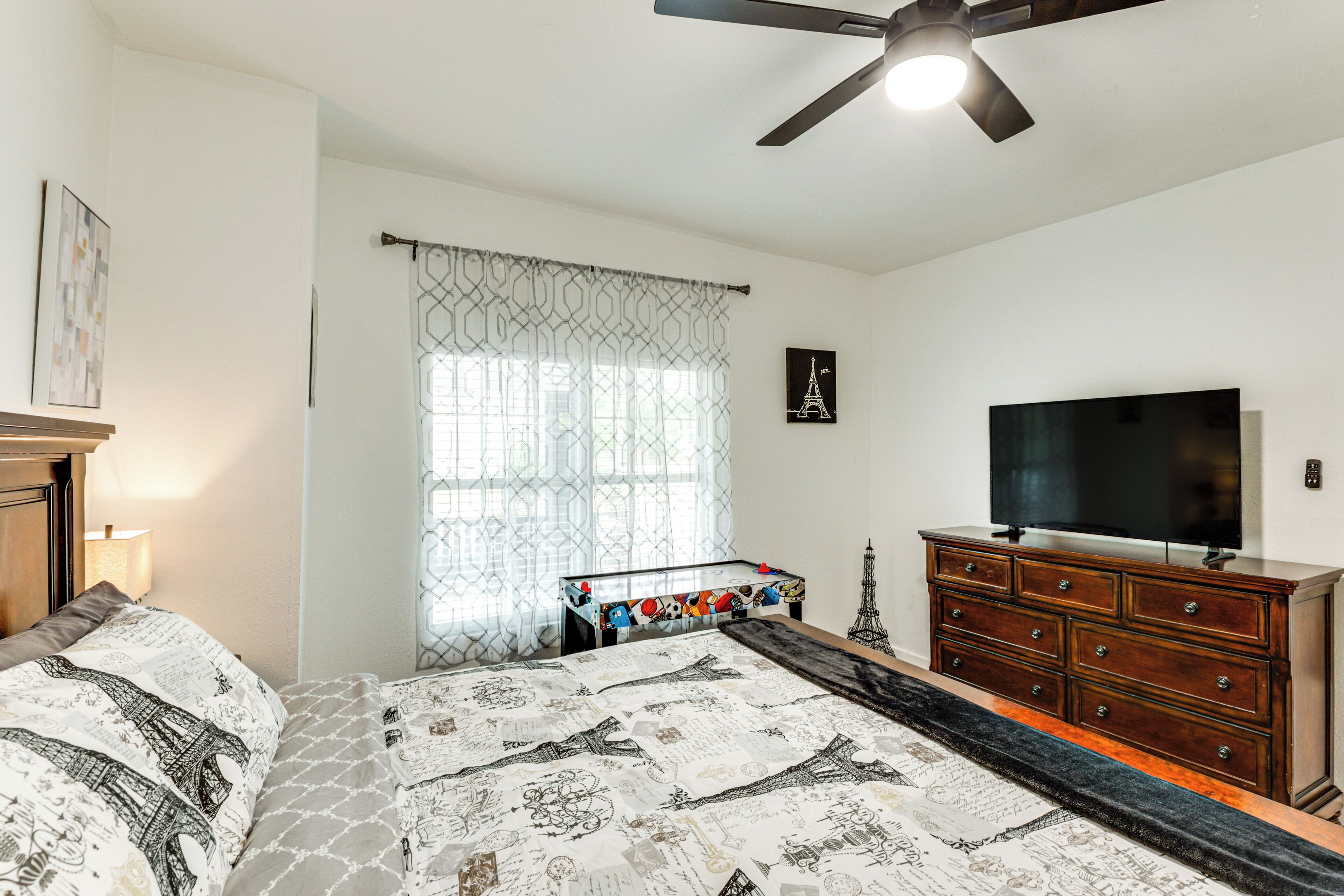 A bedroom with a bed, a dresser, a TV, a ceiling fan, and a window with patterned curtains.