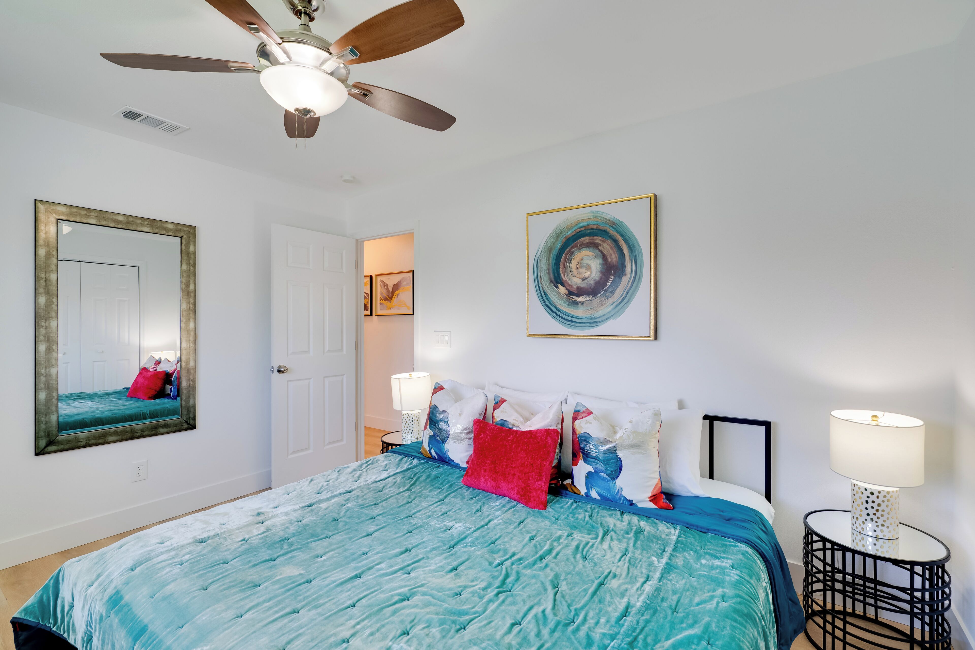 Une chambre à coucher avec un lit, un ventilateur de plafond, une table ronde avec une lampe et un tableau encadré au mur.