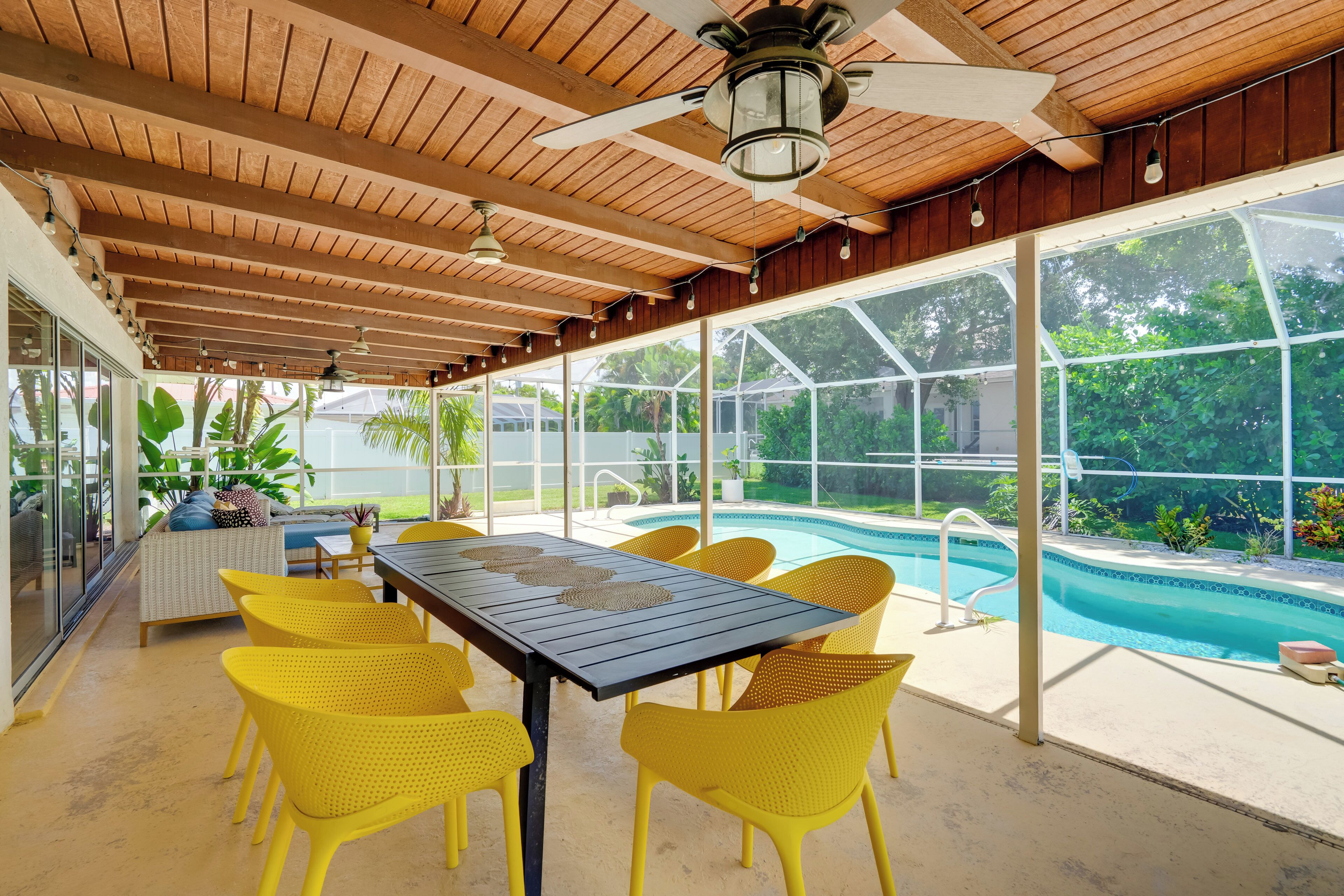 Un espace extérieur abrité avec une piscine, des chaises jaunes et une table surmontée d’un ventilateur de plafond.