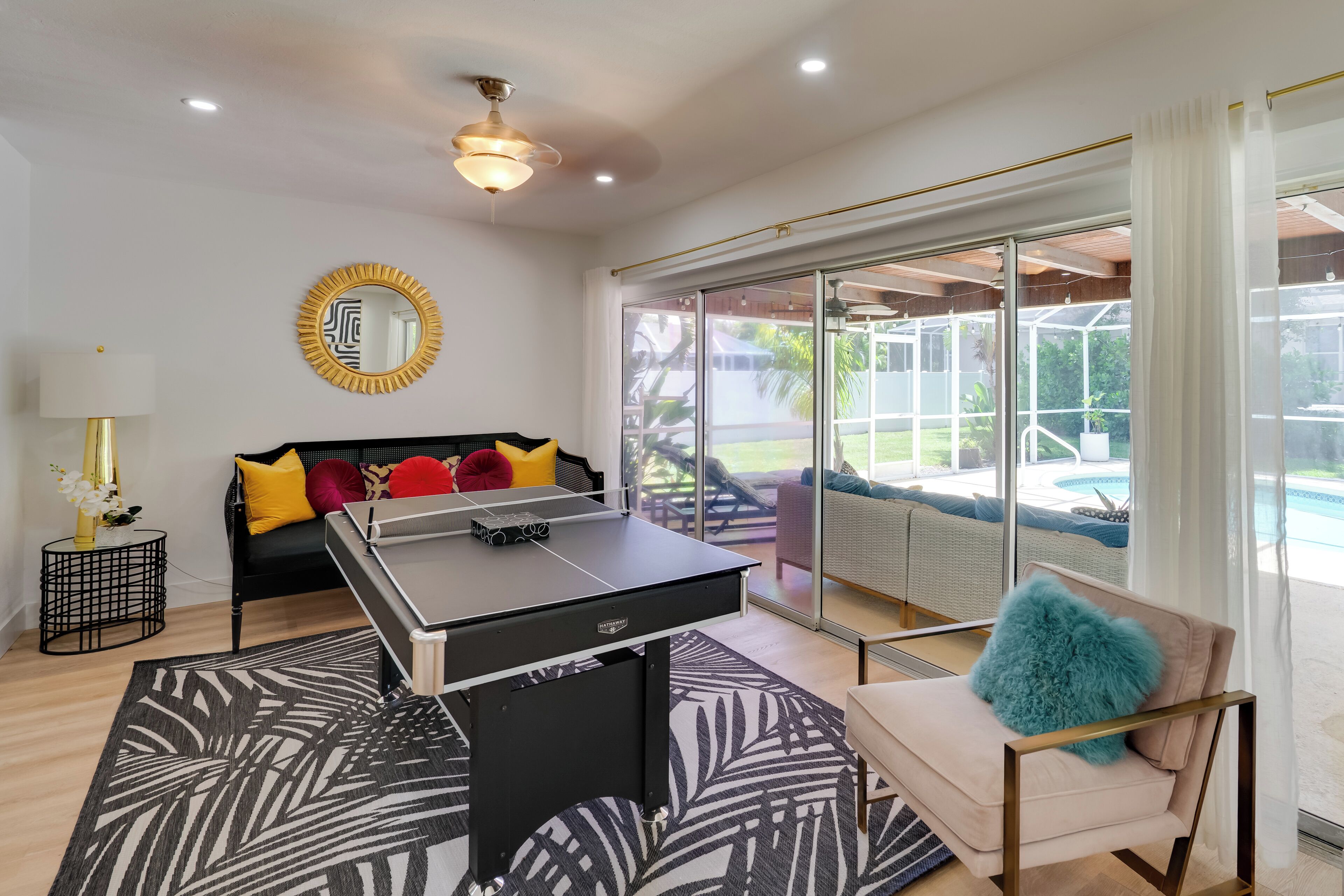 Uma sala de estar moderna com mesa de futebol, um sofá com almofadas coloridas, uma poltrona com travesseiro macio e uma grande janela com vista para uma piscina e móveis de exterior.
