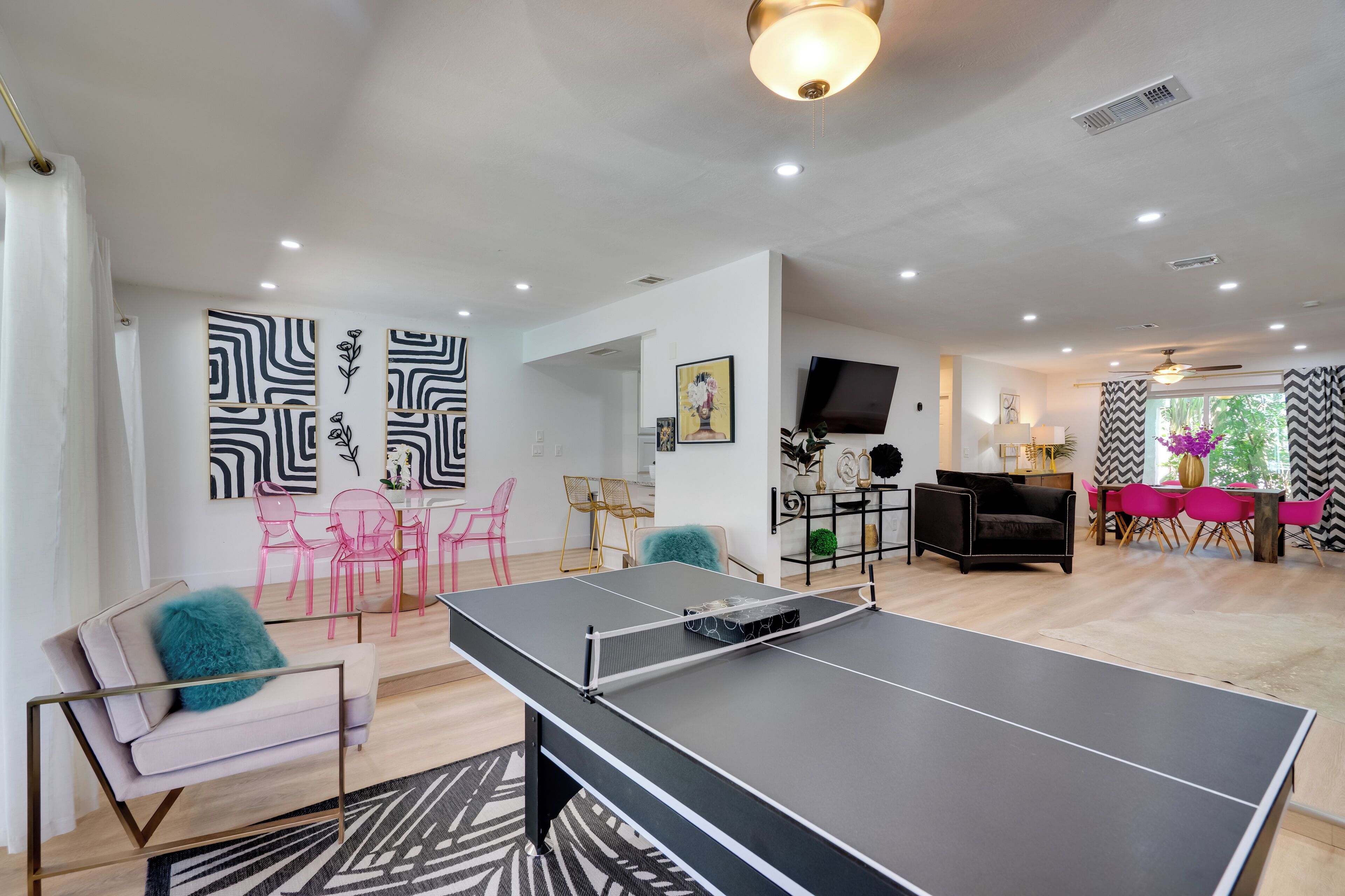 Un salon moderne avec une table de ping-pong, un canapé agrémenté d’un coussin turquoise et un coin repas avec des chaises roses.