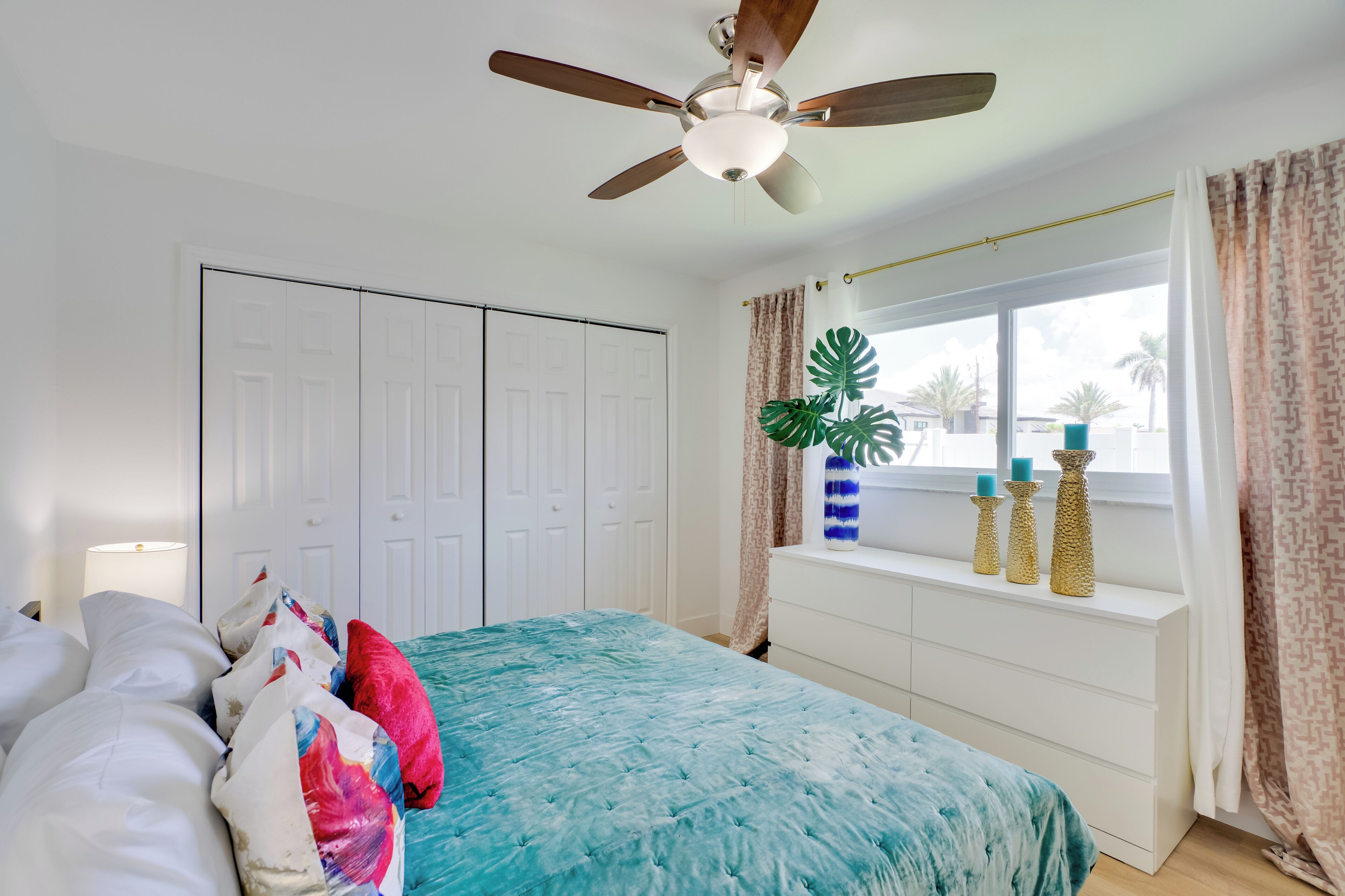 Quarto com cama, ventilador de teto, janela com cortinas e planta no parapeito.
