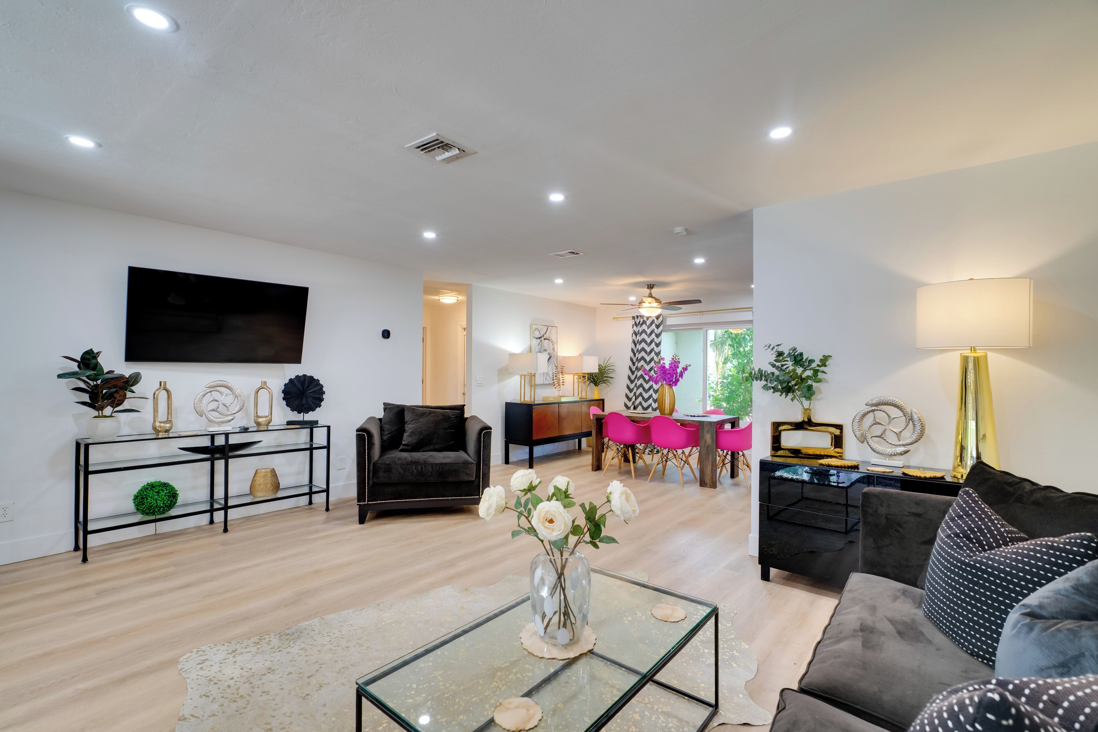 A modern living room with a glass coffee table, a sofa, a TV mounted on the wall, and a dining area with pink chairs.