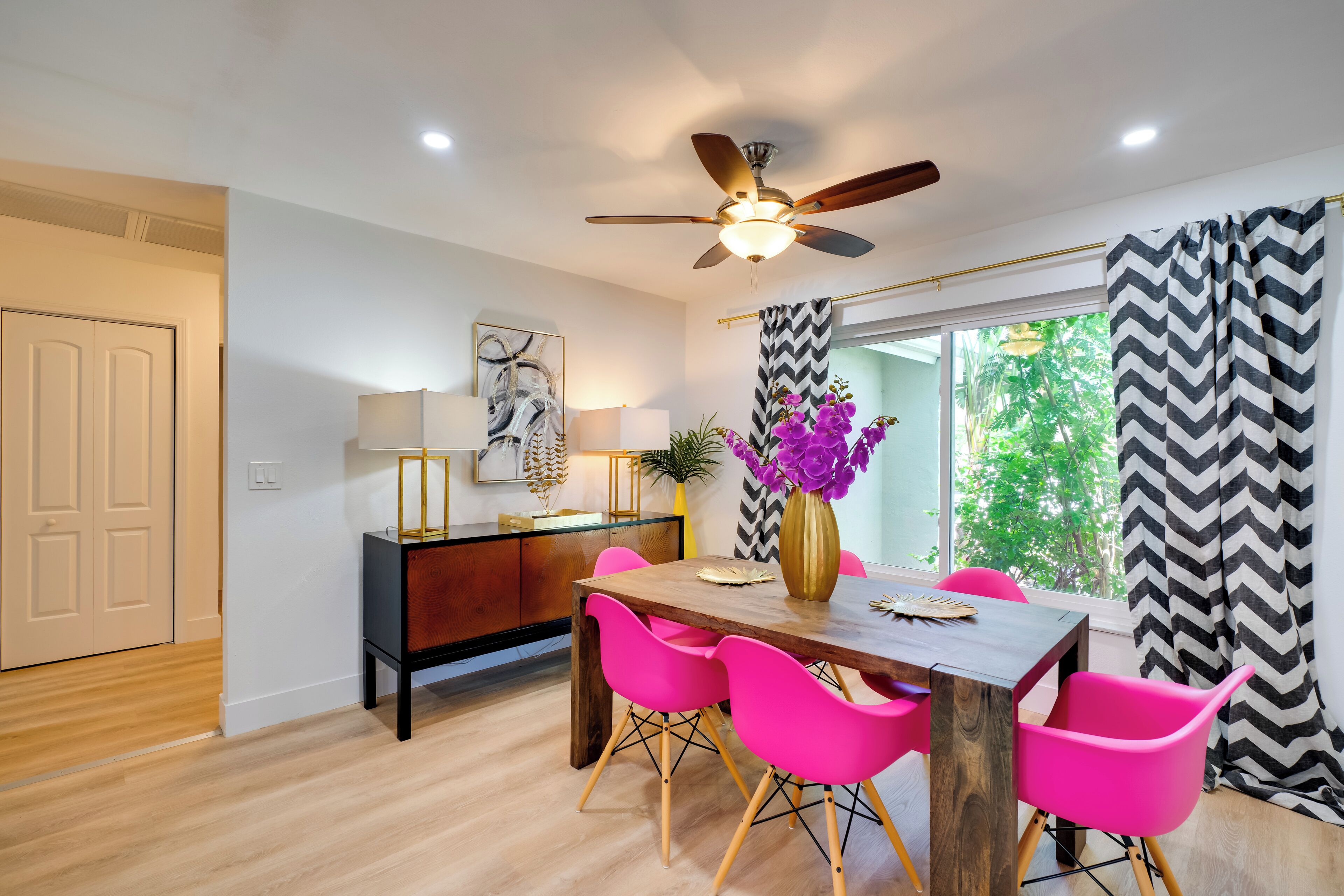 A dining area with a wooden table, pink chairs, a ceiling fan, and a window with curtains.