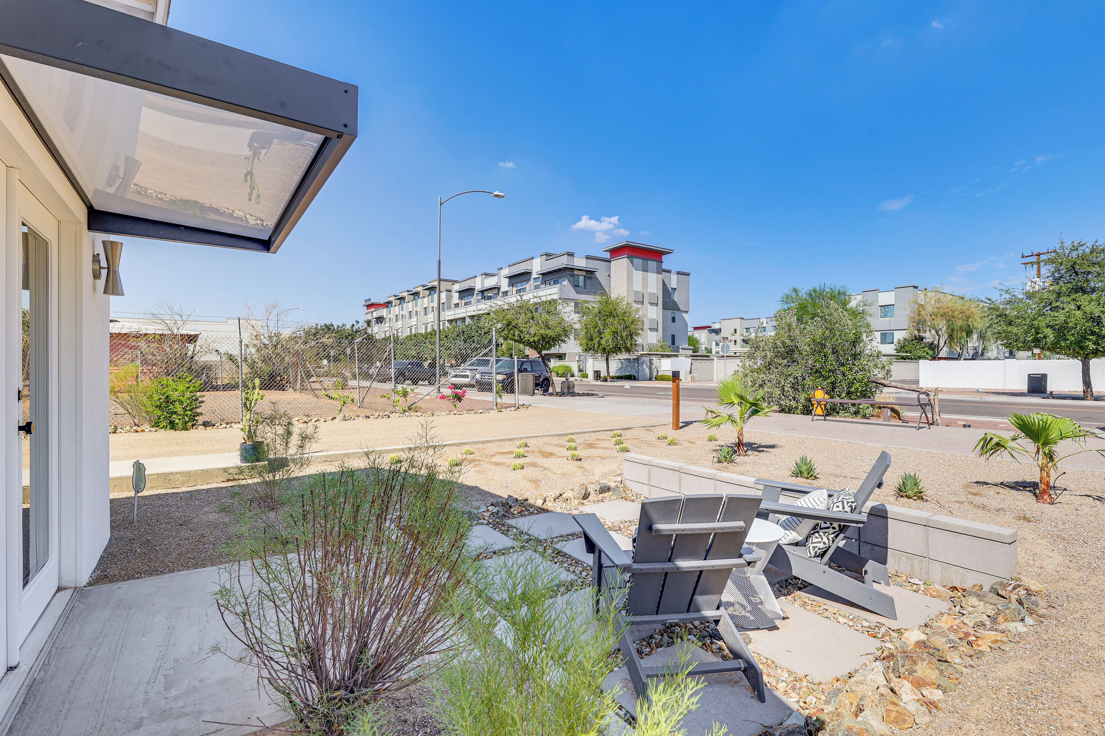 Une terrasse extérieure moderne, dotée d’un espace salon, offrant une vue dégagée sur un immeuble à plusieurs étages en arrière-plan, et agrémentée d’un jardin paysager avec différentes espèces de plantes.