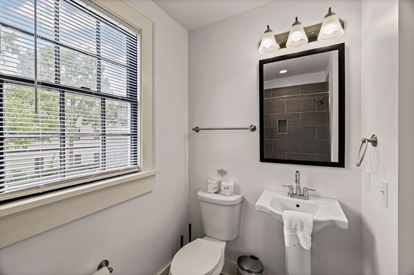A bathroom with a toilet, sink, and shower. There is a window with blinds, a towel hanging on the sink, and a mirror above the sink.