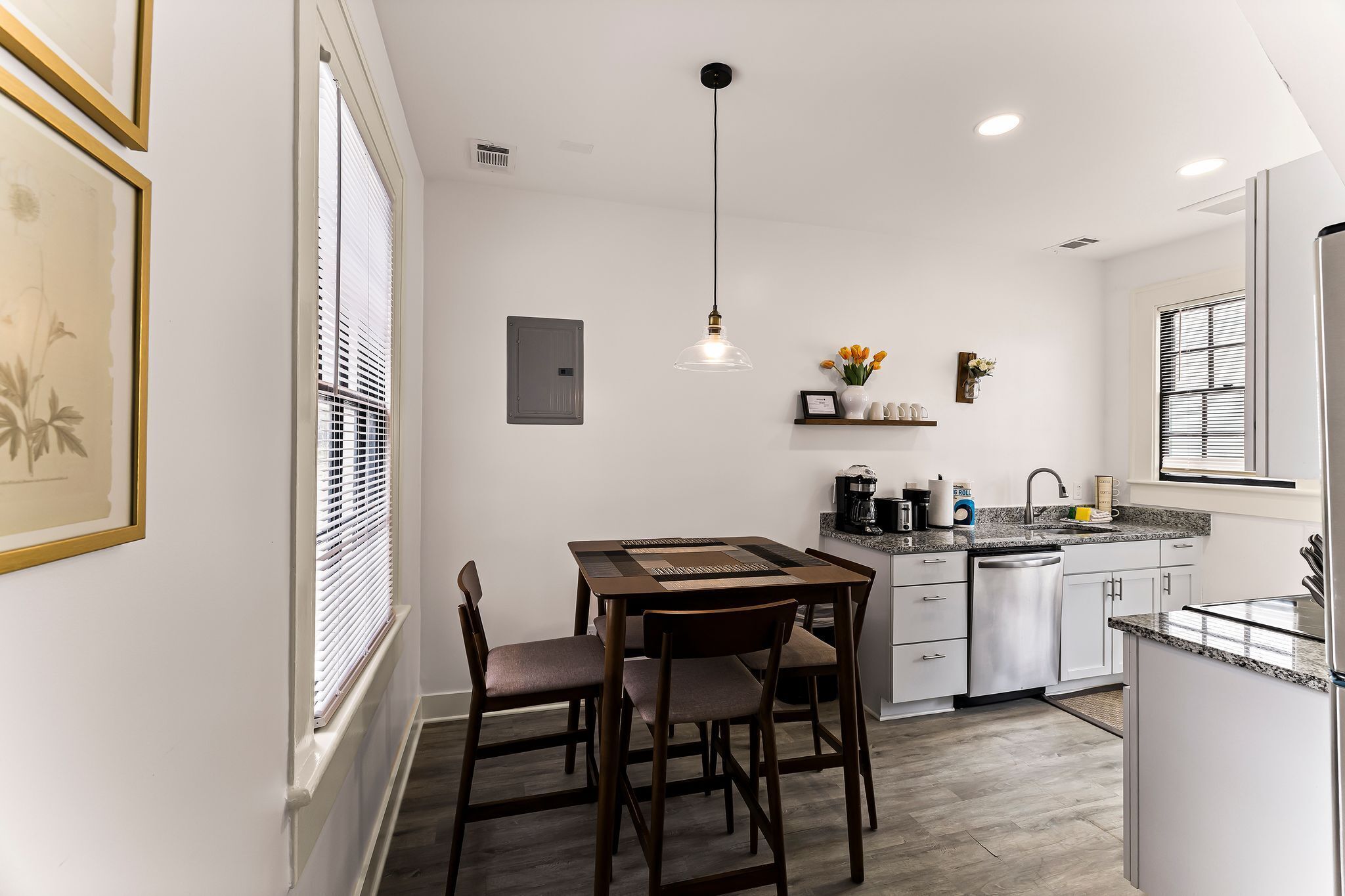 A modern kitchen with white cabinets, a dining table with chairs, a pendant light, and a window with blinds.