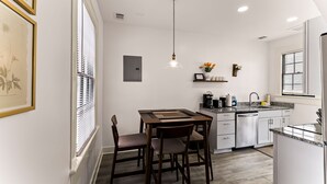 Une cuisine moderne avec des meubles blancs, une table à manger avec des chaises, un luminaire suspendu et une fenêtre avec des stores.