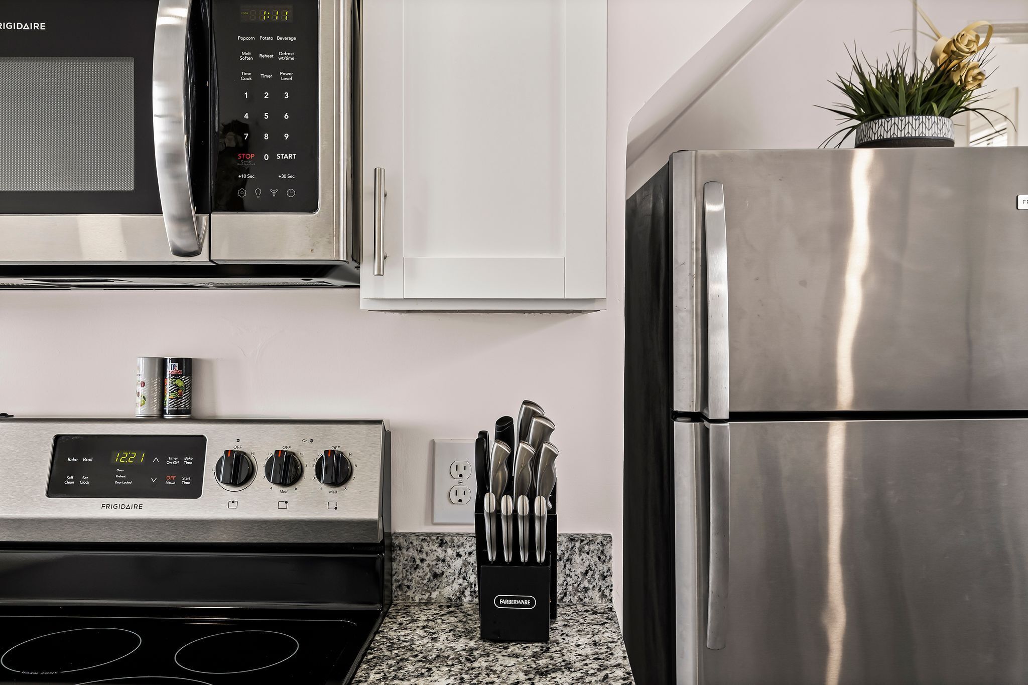 A kitchen with a microwave, oven, and refrigerator. There is a timer on the oven and a knife block on the counter.