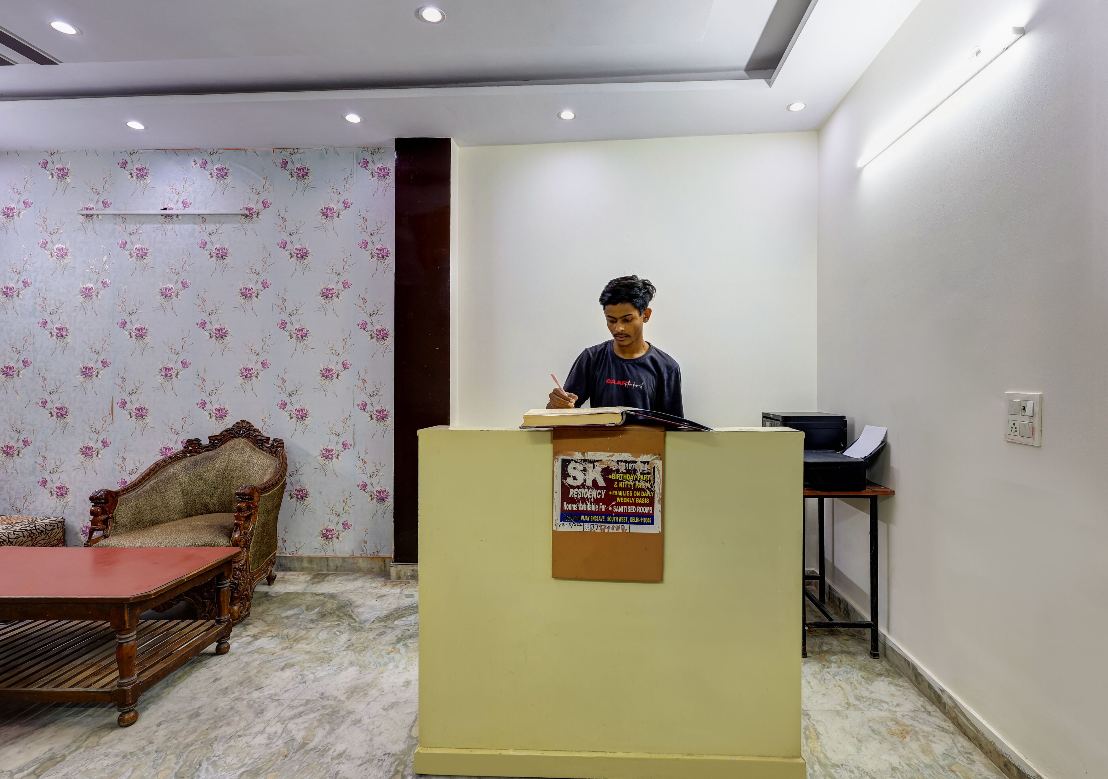 A room with a wooden sofa, a small table, a desk with a printer, and a bulletin board with a notice.