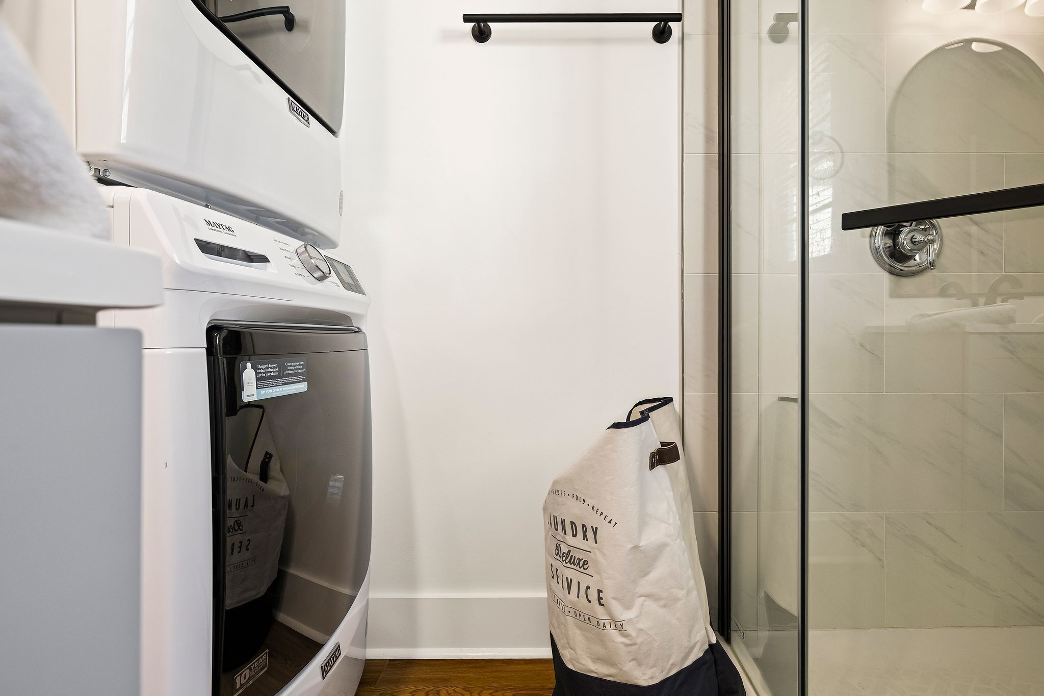 A laundry bag hanging near a glass shower door, a white washing machine, and a white dryer in a bathroom.