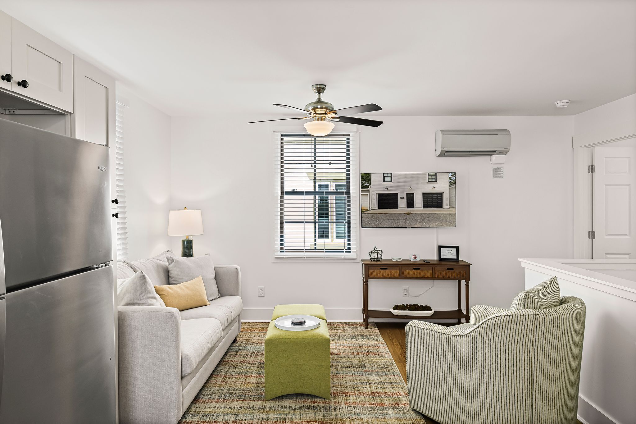A compact living space with a kitchenette, a ceiling fan, a window with blinds, a sofa, a small ottoman, a side table, and a striped chair.