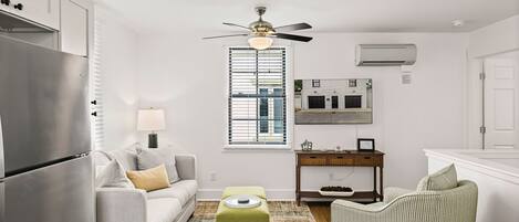 A compact living space with a kitchenette, a ceiling fan, a window with blinds, a sofa, a small ottoman, a side table, and a striped chair.