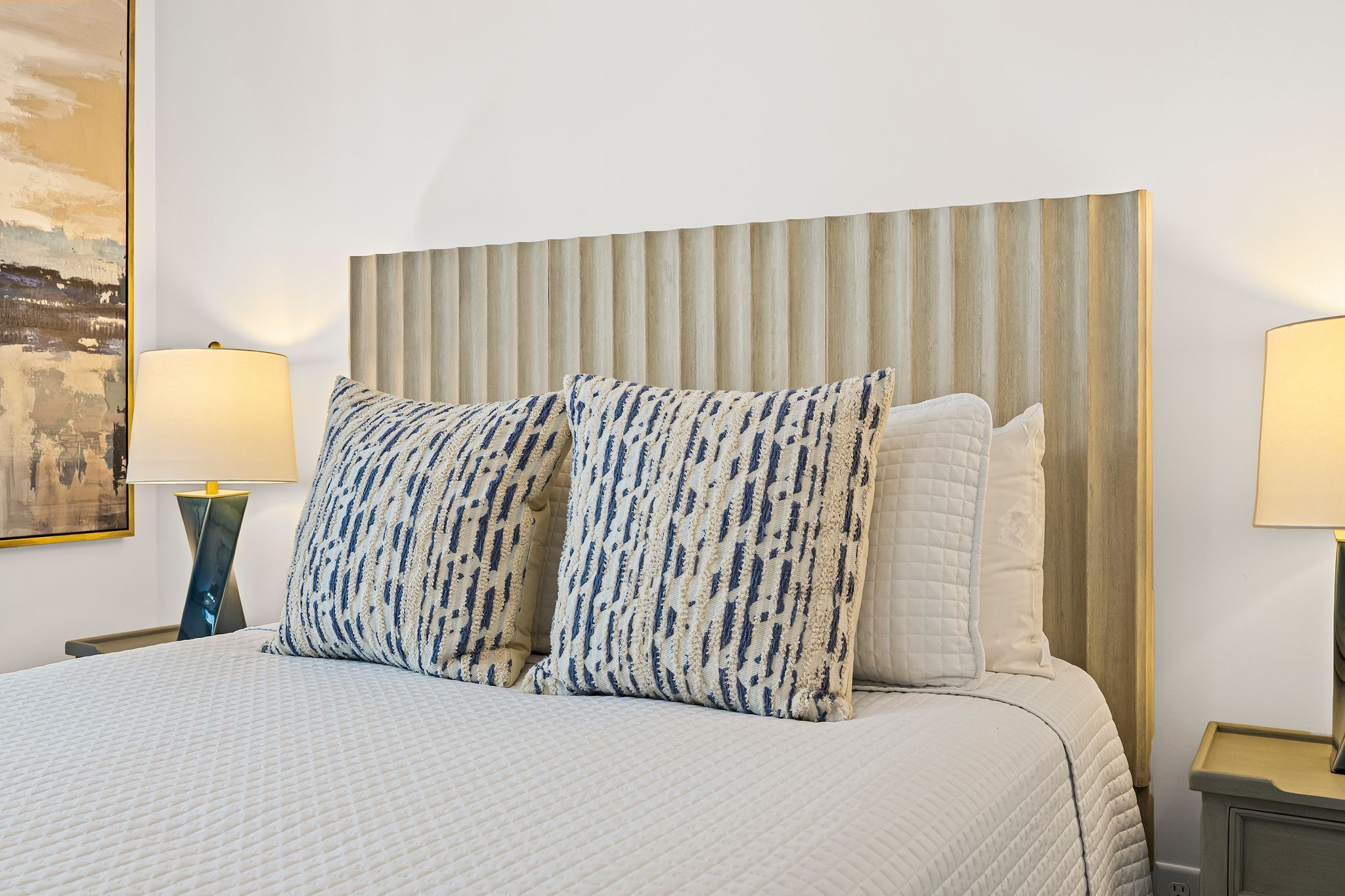 A neatly made bed with a patterned pillow and a quilted blanket, flanked by two bedside lamps and a wall-mounted painting.
