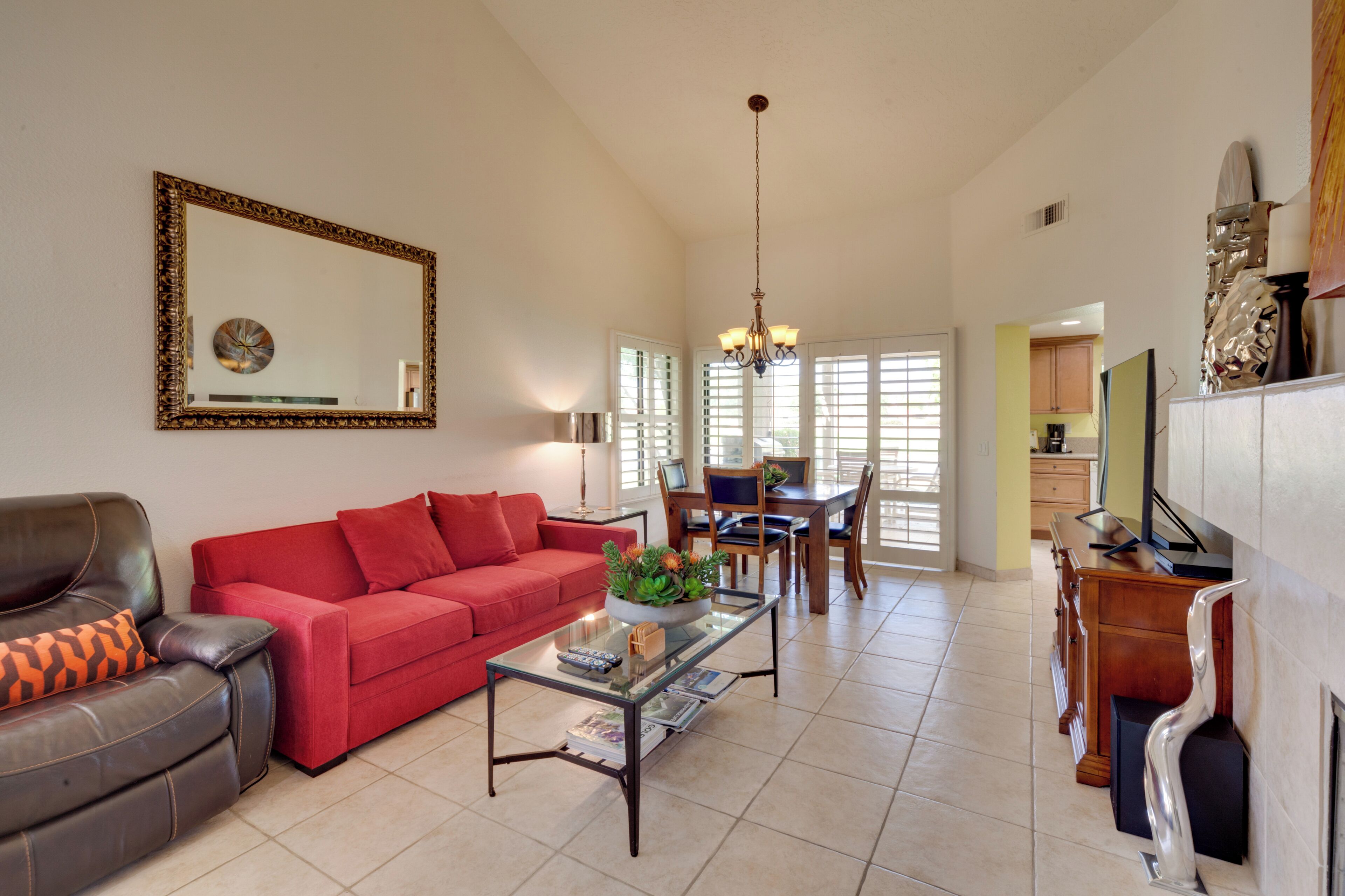 A spacious living room with a red sofa, a leather armchair, a glass coffee table, and a dining area with wooden chairs and a chandelier.