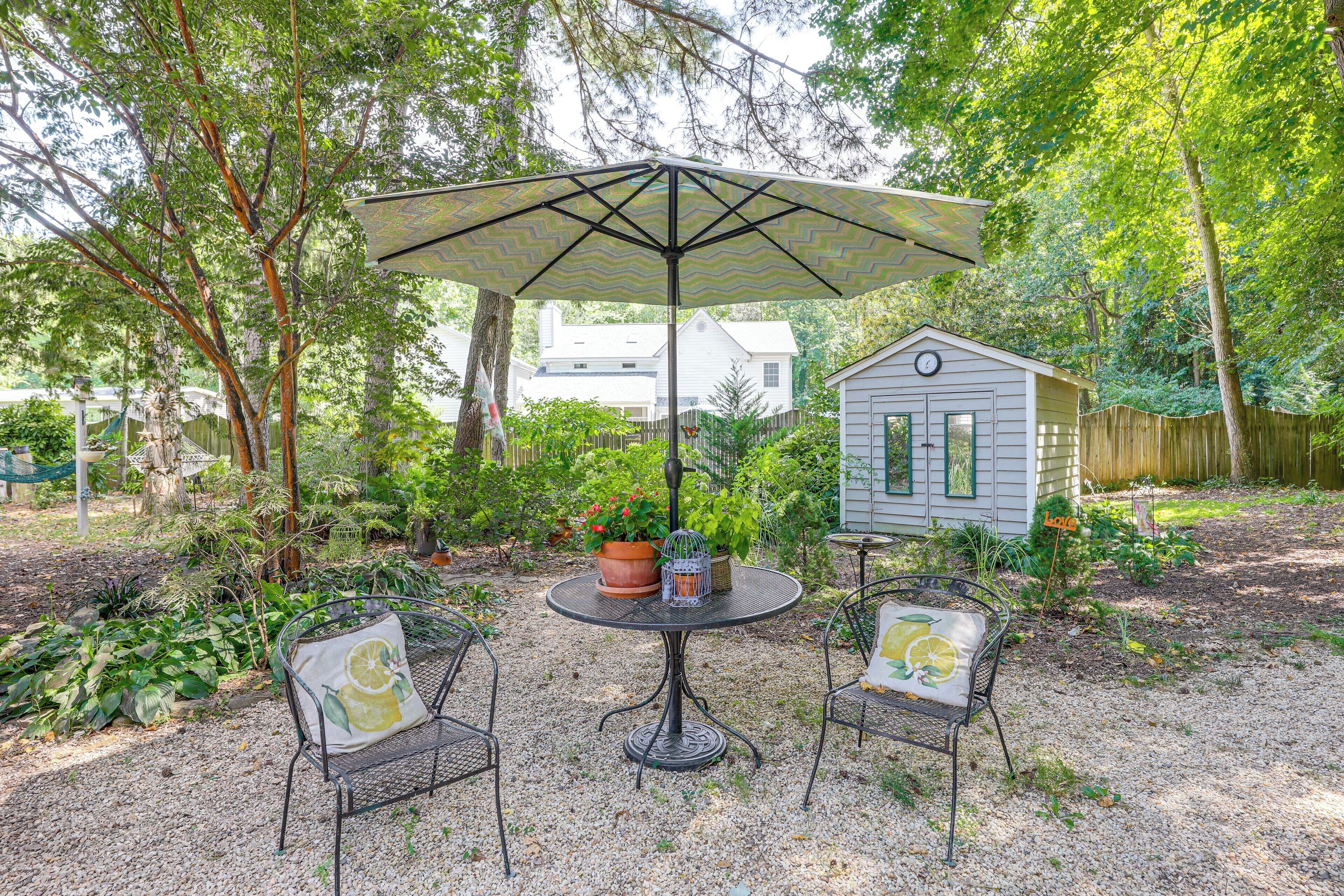 A patio with a round table and two chairs, an umbrella, and a small shed in a garden setting.
