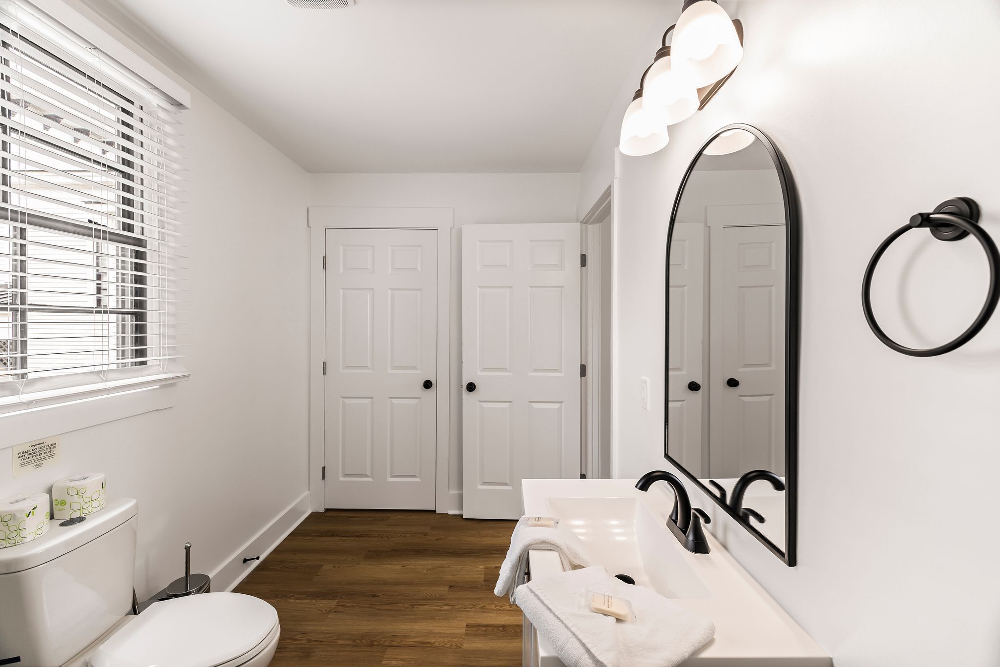 Baño con lavamanos blanco, grifo negro y un espejo grande. Se observan dos puertas blancas al fondo y una ventana con persianas.