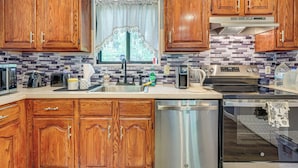 A kitchen with wooden cabinets, a stainless steel dishwasher, a built-in oven, and a window with a curtain. - Poconos Family Home w/ Game Room + Community Perks (East Stroudsburg)