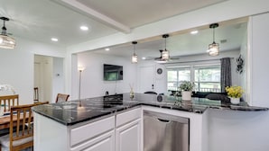 A modern kitchen with white cabinetry, a black marble countertop, stainless steel dishwasher, pendant lighting, and a dining area with wooden chairs.
