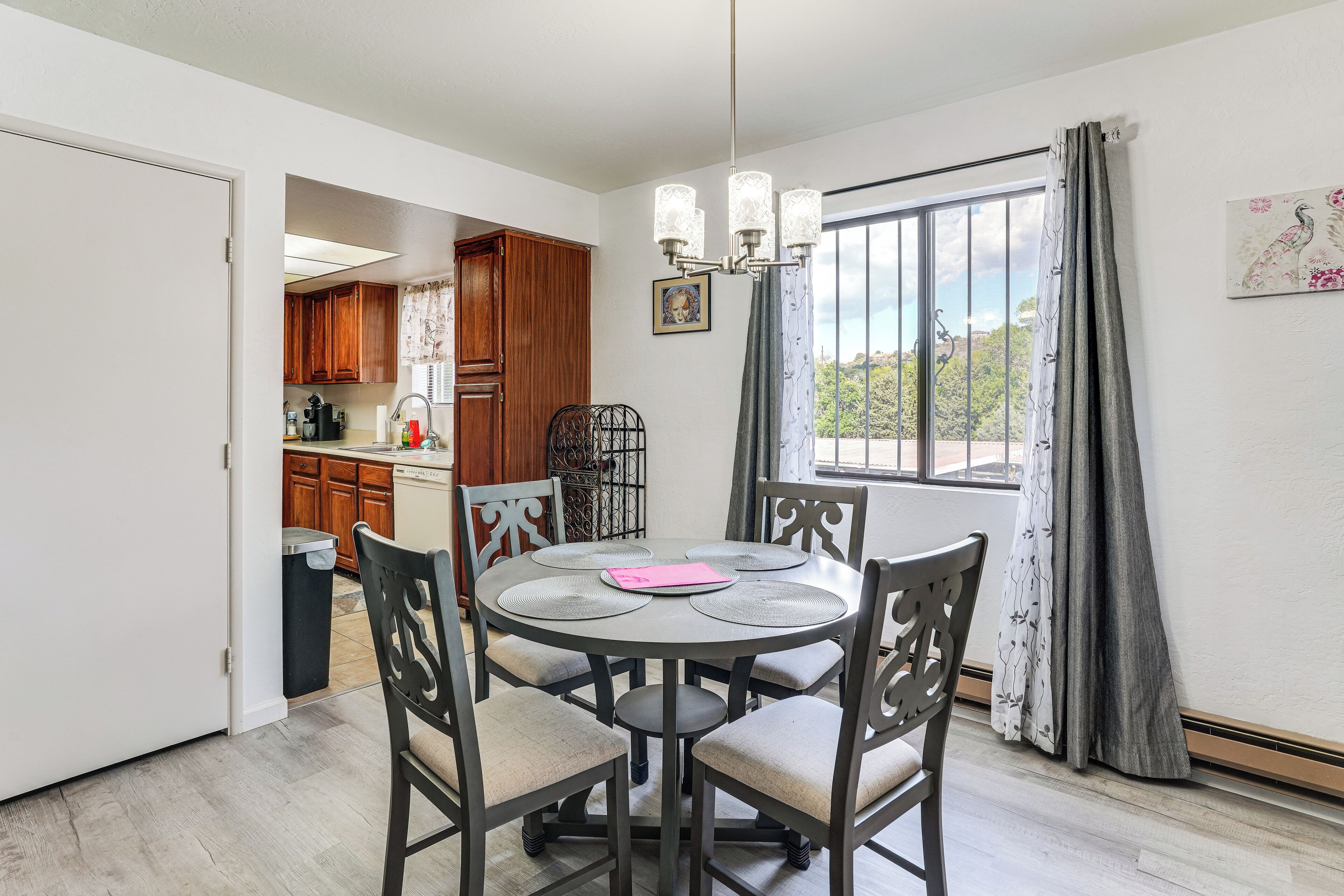 A dining area with a round table and chairs, a chandelier, and a view of the outdoors through a window with curtains.