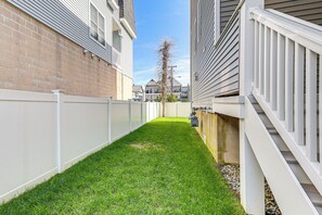 Un patio trasero estrecho y cubierto de césped, con una cerca blanca, un muro de hormigón y una escalera que sube a un edificio con barandilla blanca.