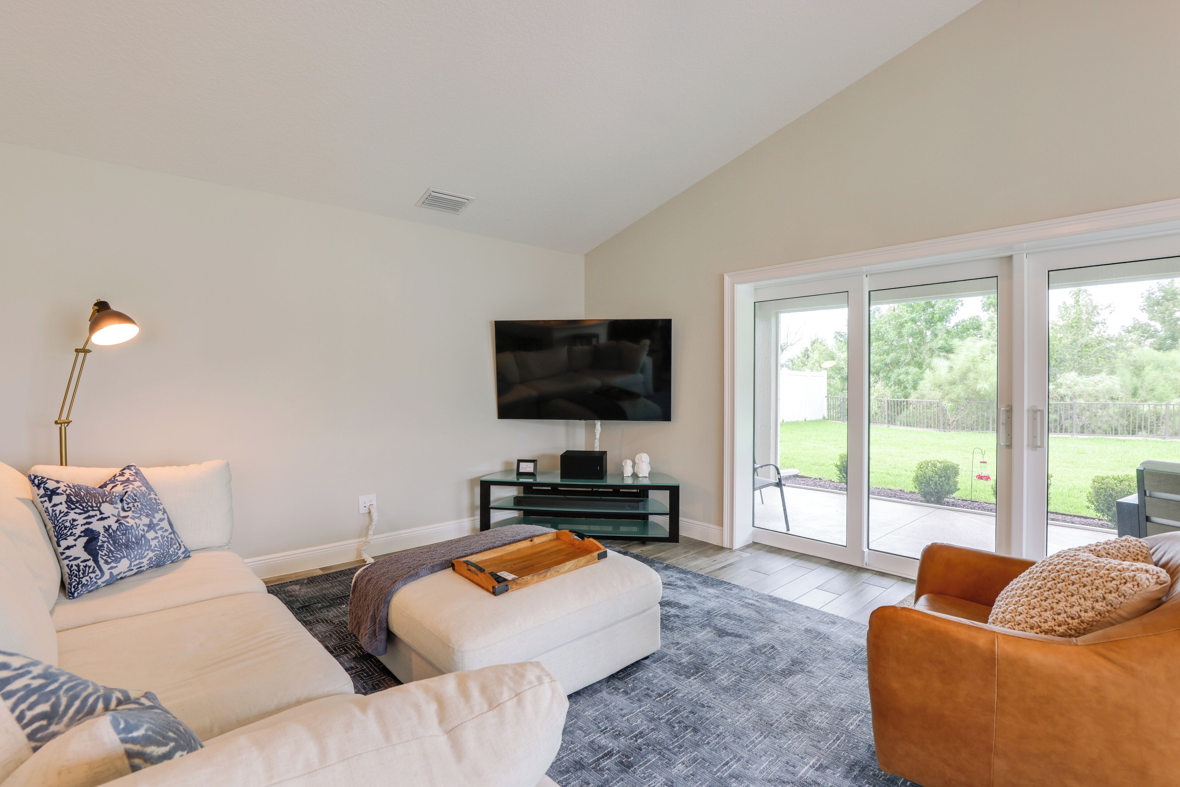 A living room with a sofa, ottoman, and armchair, a flat-screen TV on a stand, and a sliding glass door leading to a patio.