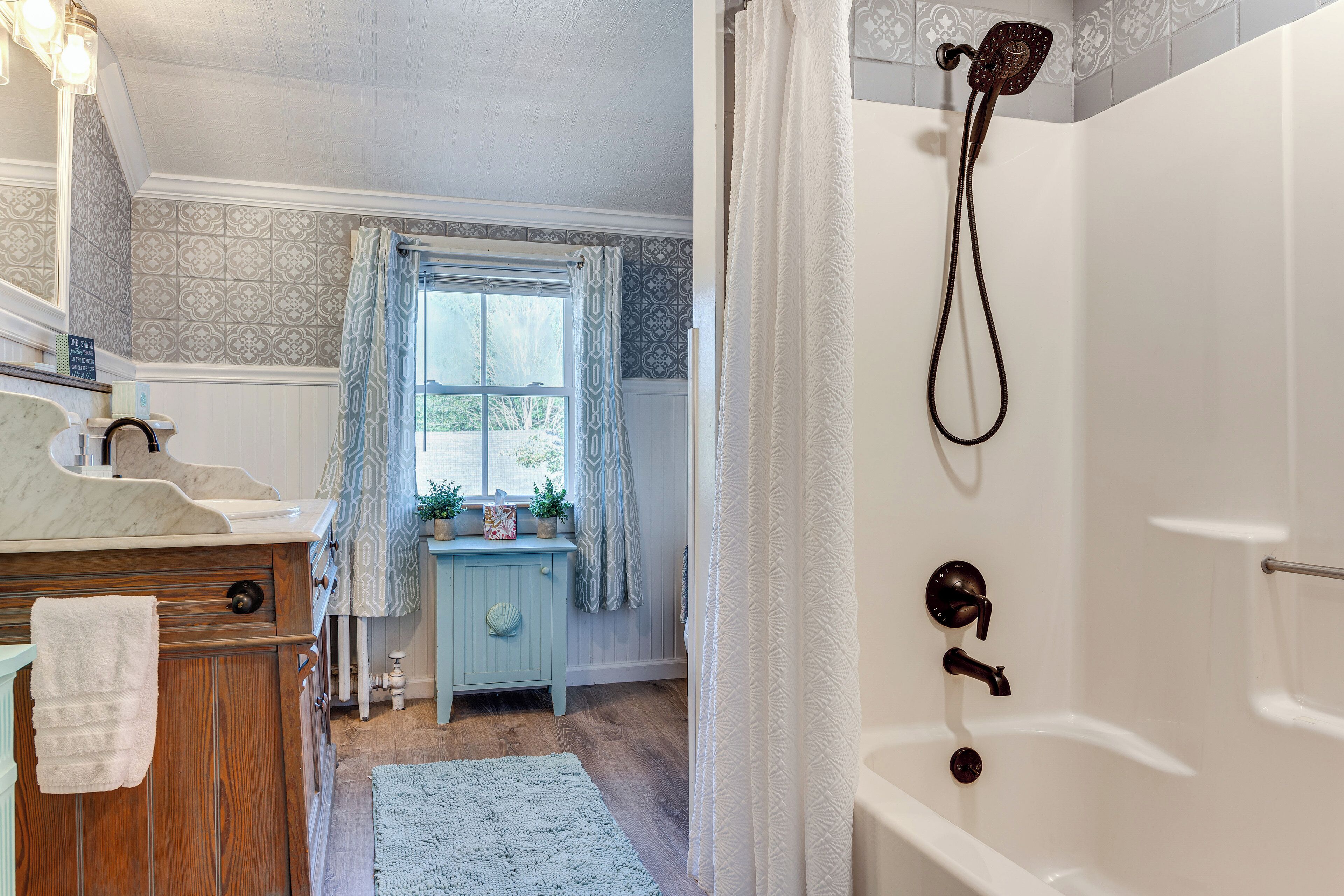 A bathroom with a bathtub, shower, vanity with a mirror, and a window with patterned curtains.