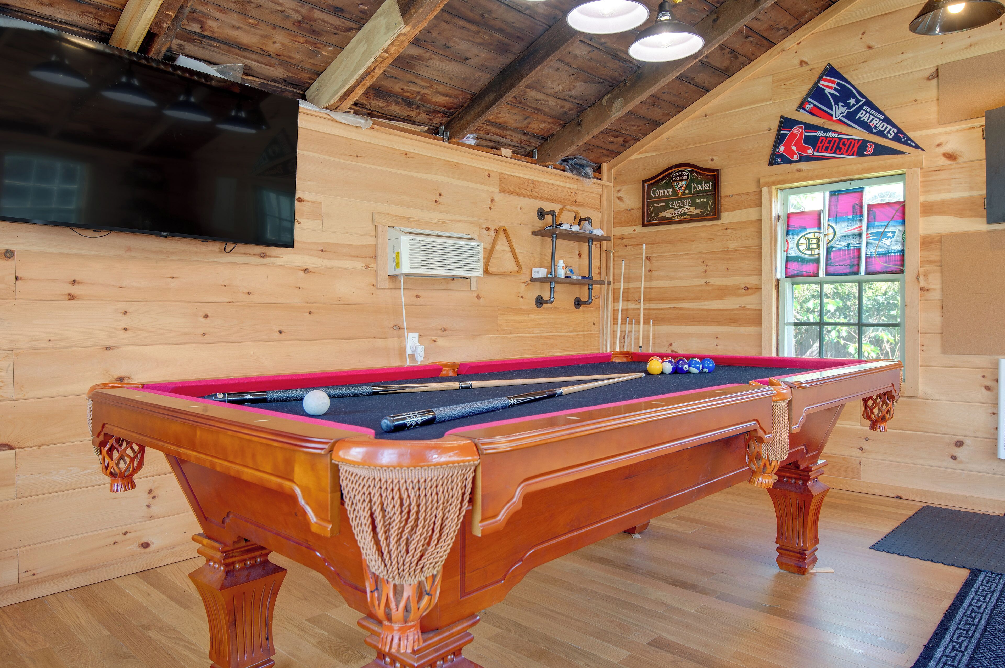 Une table de billard se trouve dans une pièce en bois, avec un téléviseur au-dessus et une fenêtre donnant sur l’extérieur.