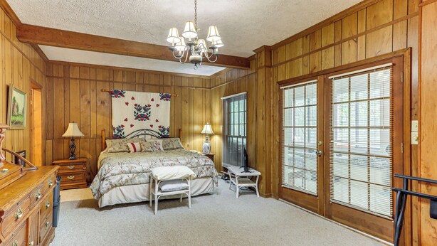 A bedroom with wooden paneling, a bed with floral bedding, a chandelier, a dresser, and a large window with blinds. - Creekside Family Home in Cleveland: Stay & Play! (Cleveland)