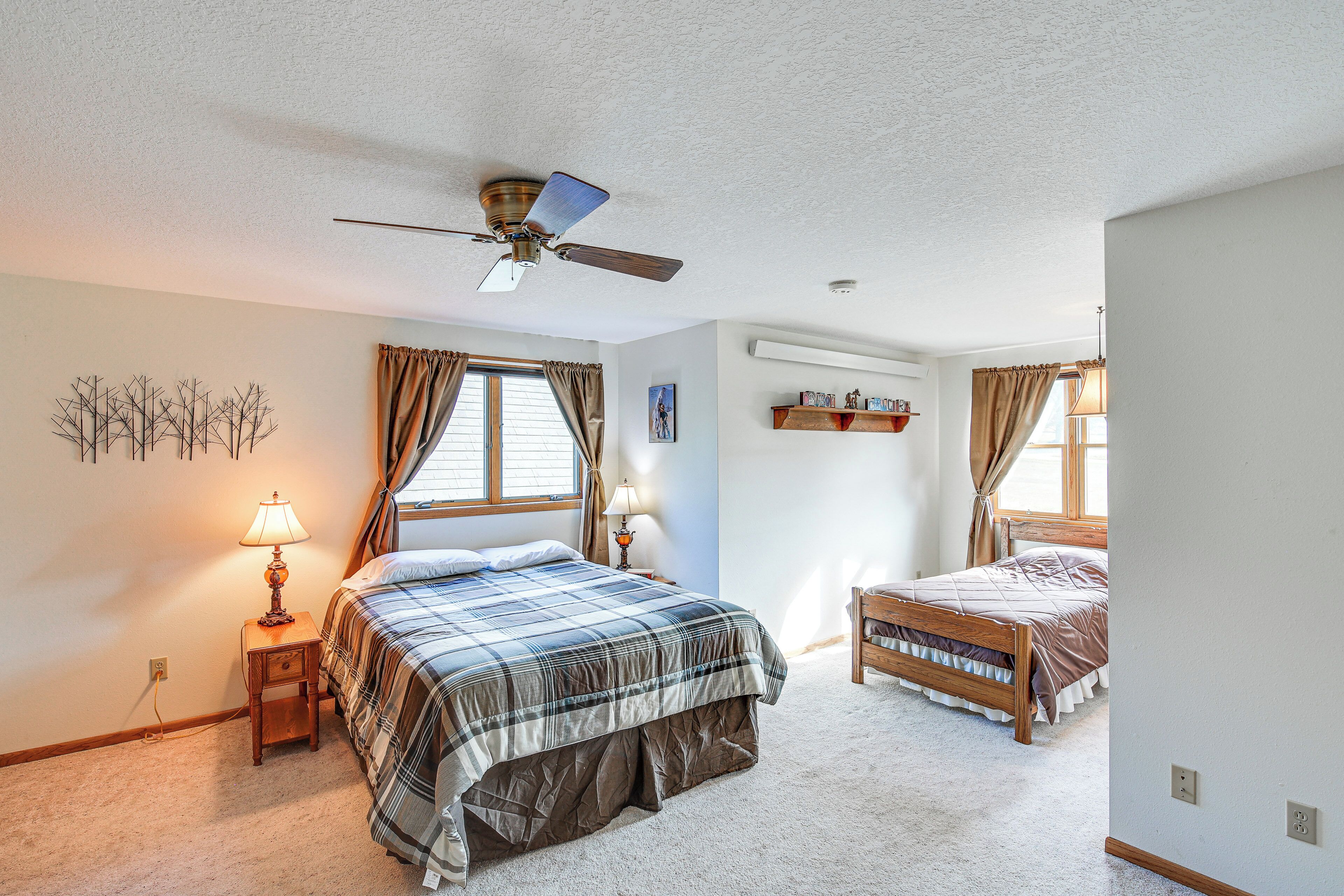 A bedroom with a large bed, a smaller bed, a ceiling fan, a nightstand with a lamp, and a window with curtains.