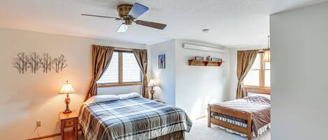 A bedroom with a large bed, a smaller bed, a ceiling fan, a nightstand with a lamp, and a window with curtains.