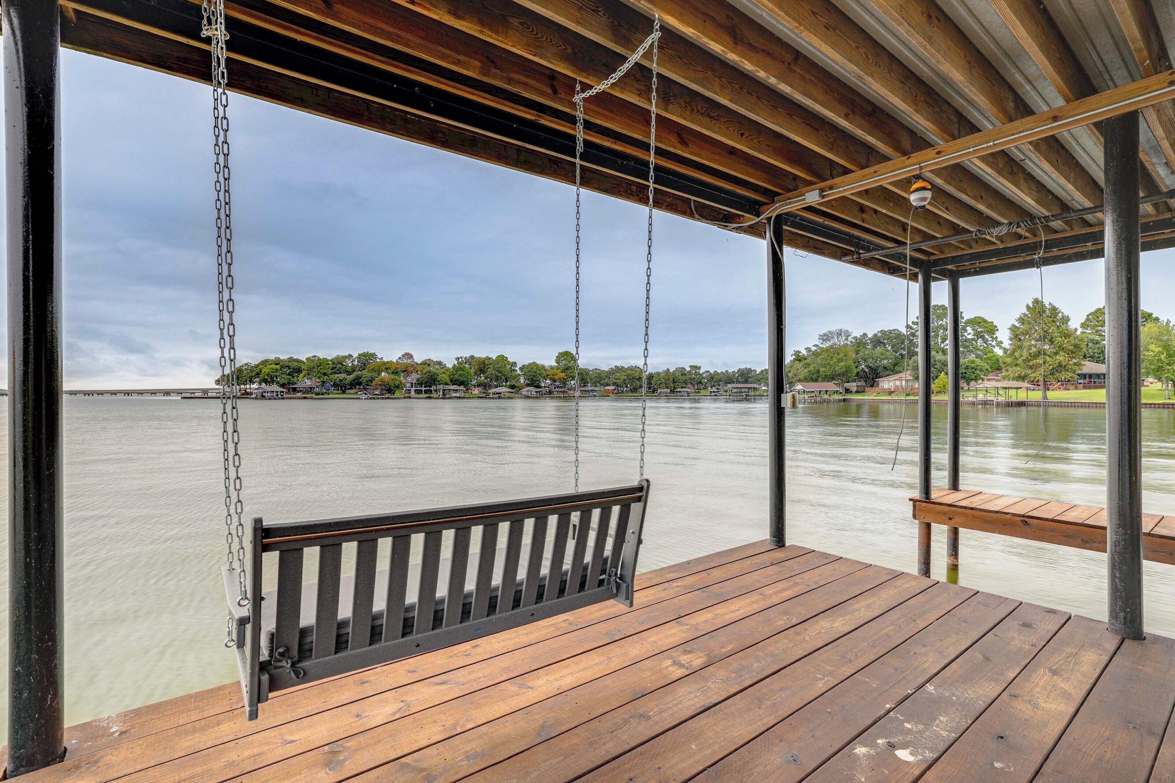 A wooden deck with a bench and a swing overlooking a calm body of water, with trees and buildings in the background.