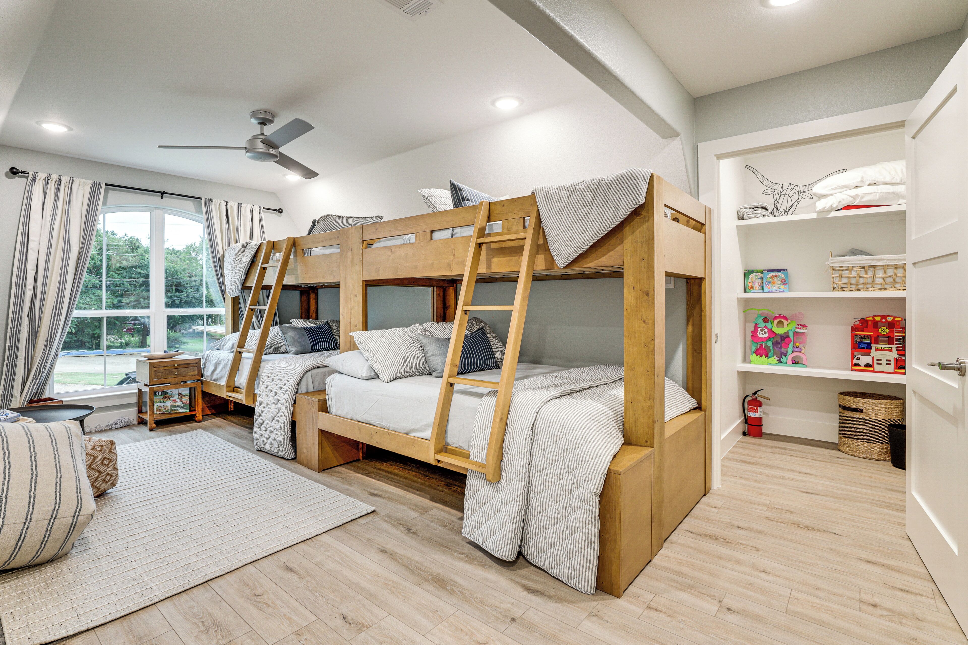 Une chambre avec un lit superposé, une échelle, un ventilateur de plafond, une fenêtre avec des rideaux et une bibliothèque remplie de jouets.