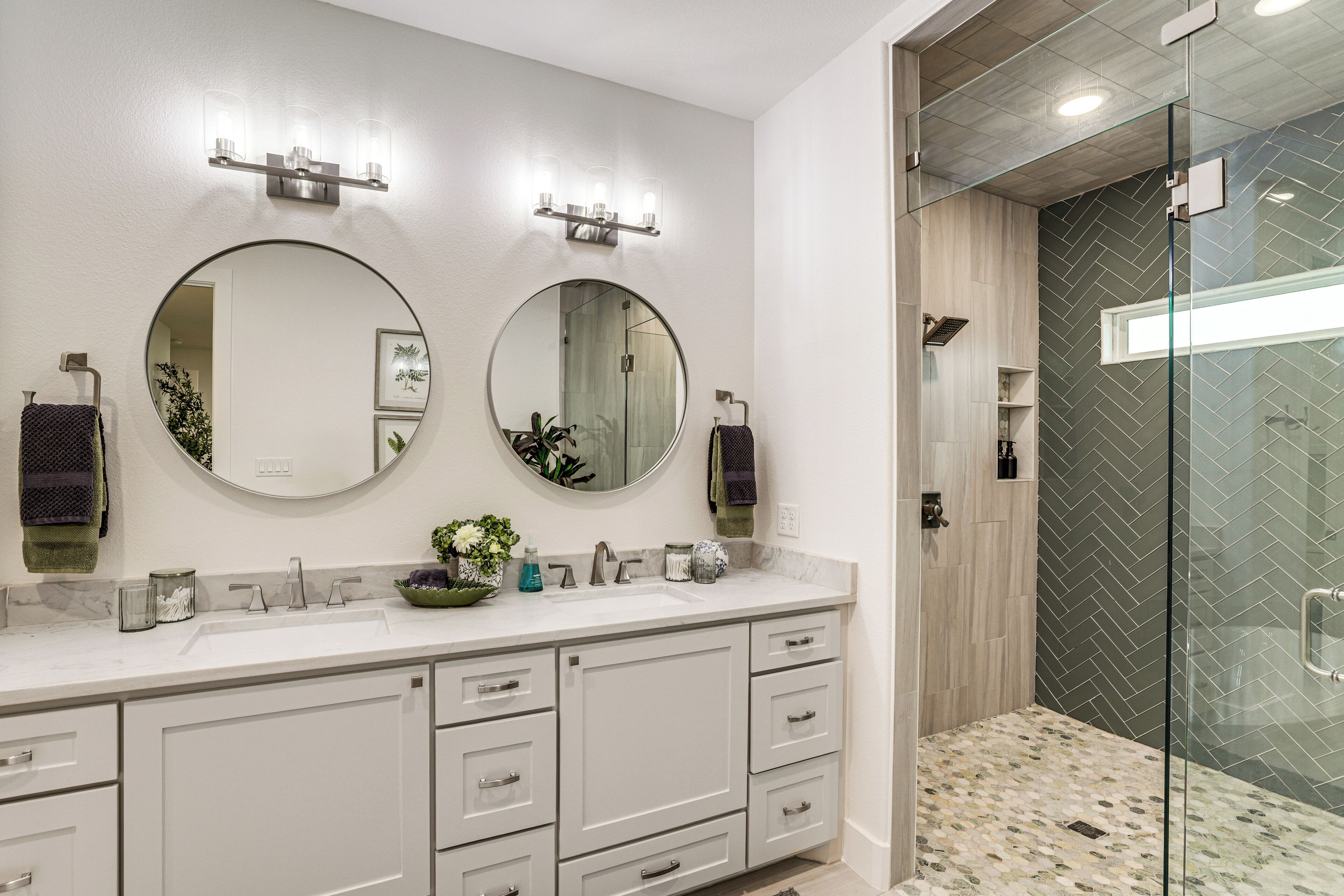A modern bathroom with two round mirrors, a white vanity with drawers, a glass shower enclosure, and a herringbone-patterned tile backsplash.