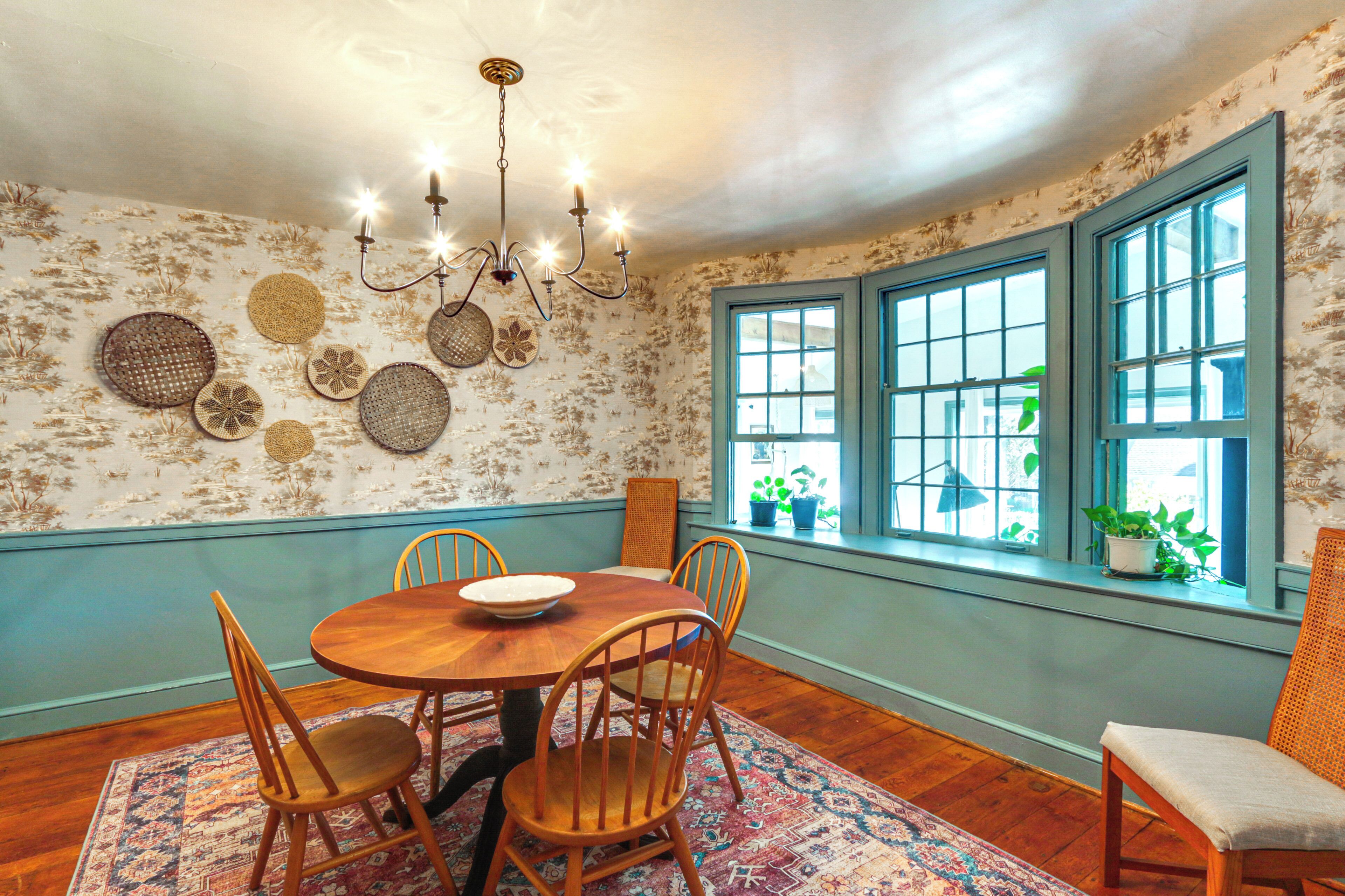 A dining room with a wooden table, four chairs, a chandelier, and a window with plants.