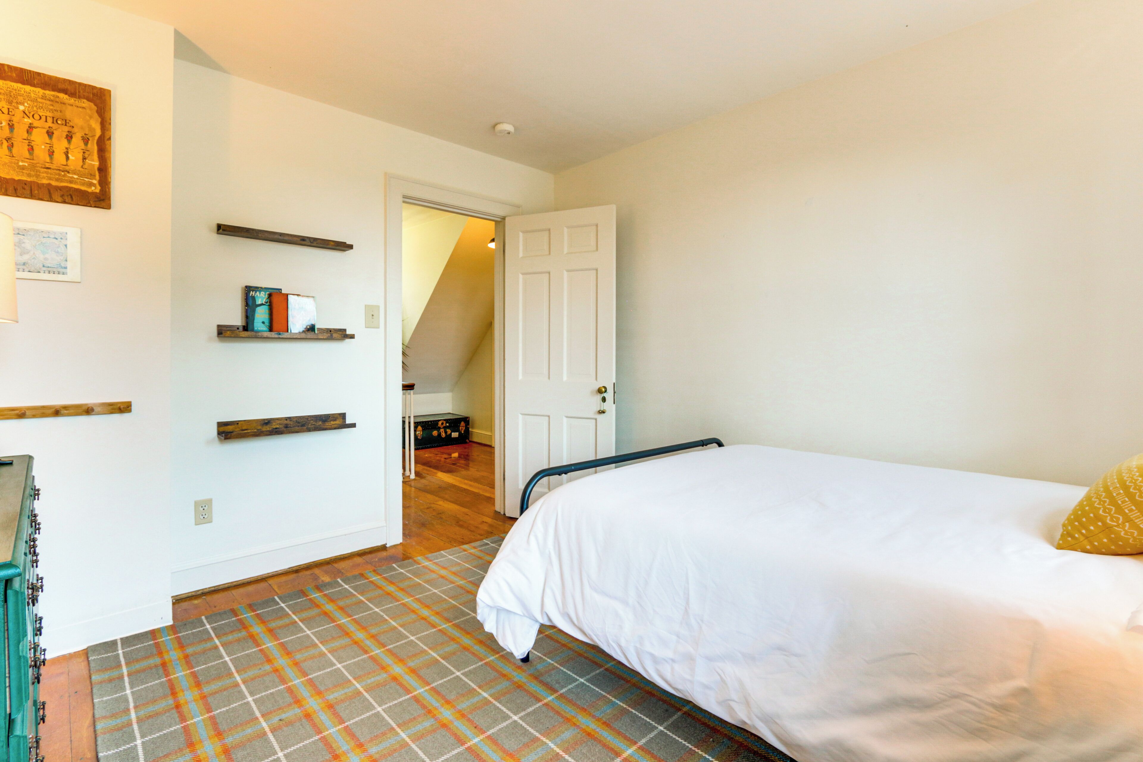 Een slaapkamer met een bed, een houten dressoir, een ruiterig tapijt en een deur naar een andere kamer.